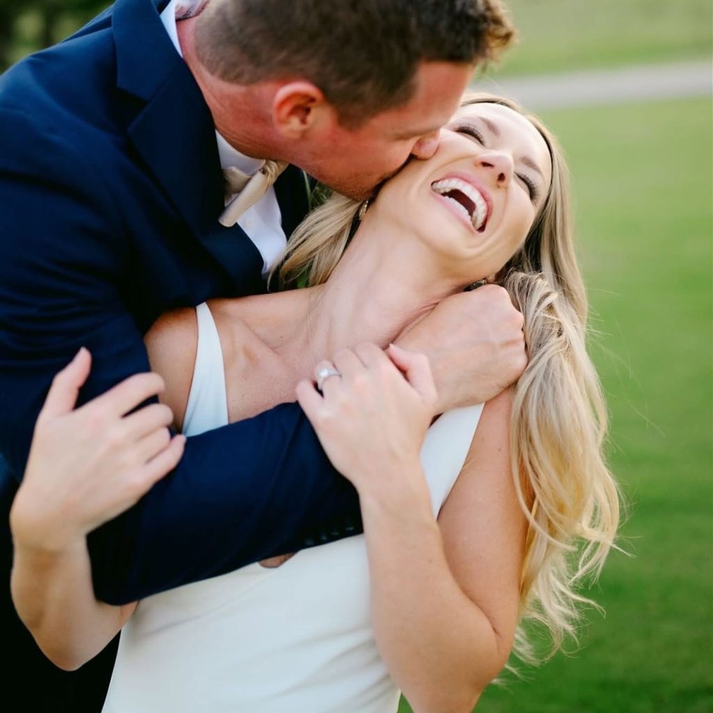 A laughing bride with loose blonde waves embraced by her groom